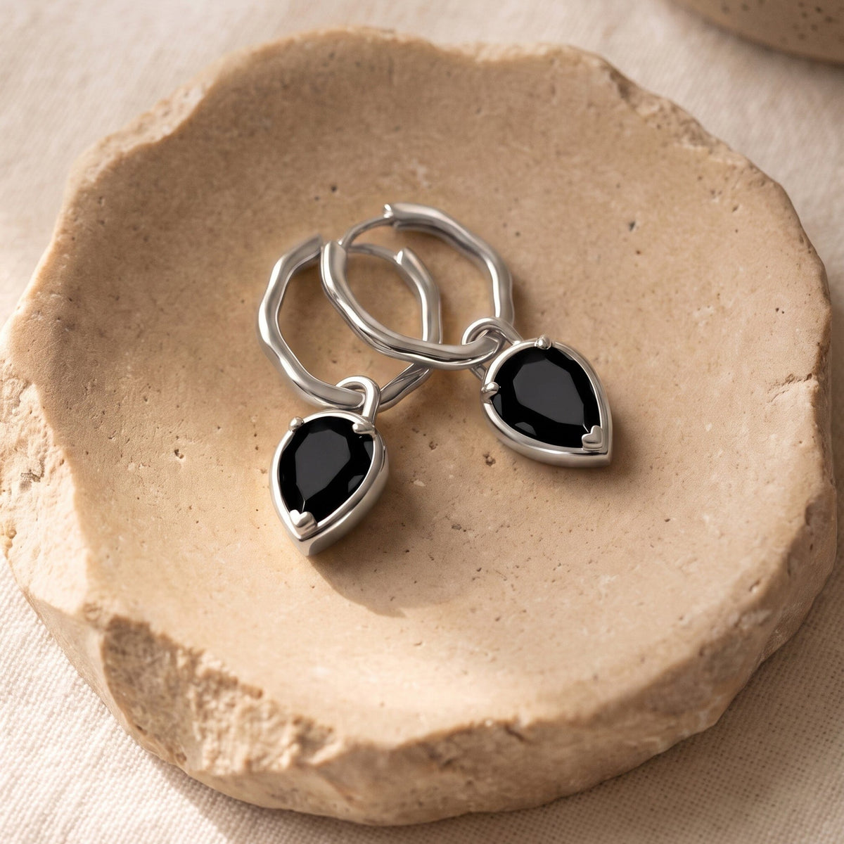 Pair of black pear-shaped stone hoop earrings arranged on a textured beige stone slab.