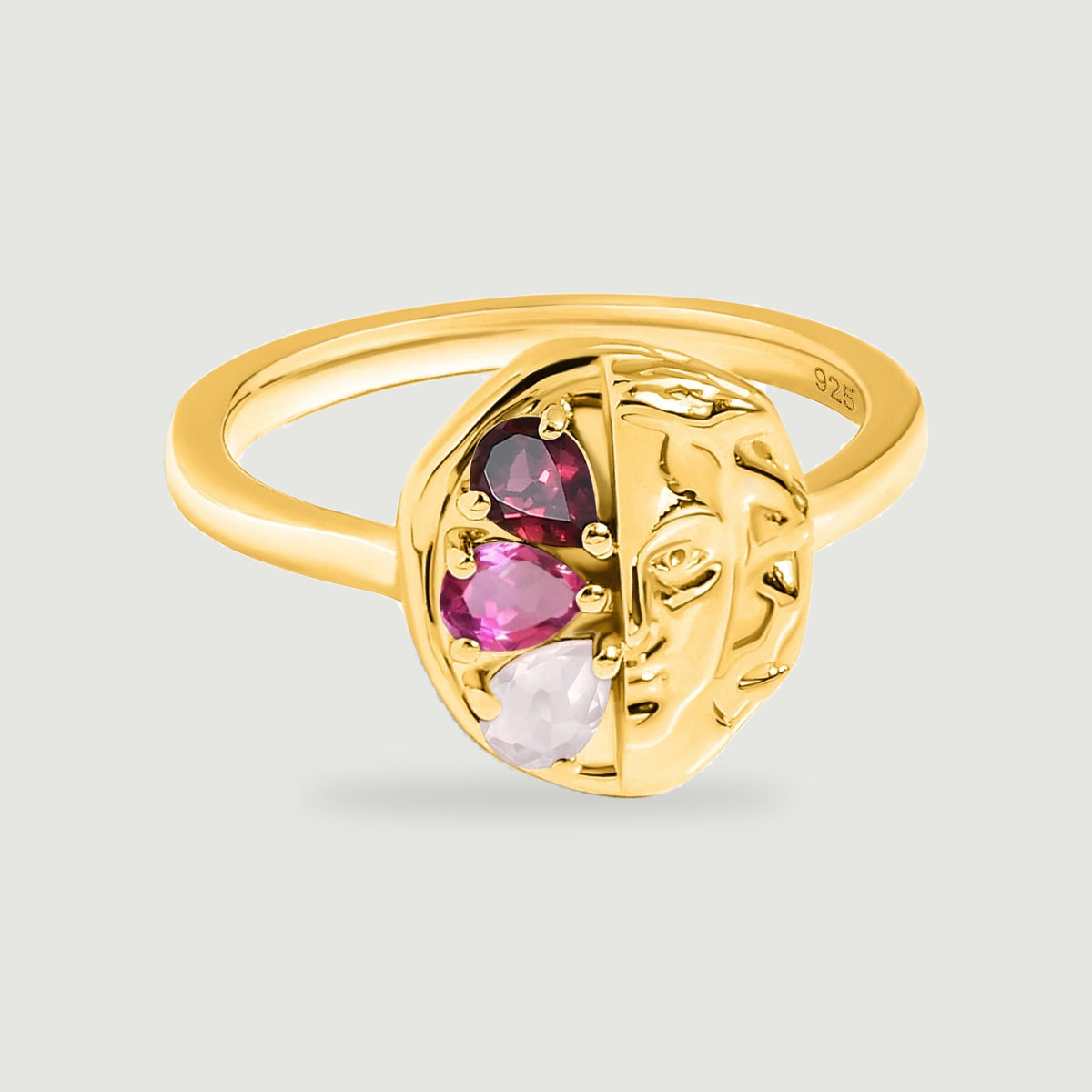 A yellow gold ring with a sculpted face and three red and pink pear-shaped stones rests on a light grey surface.