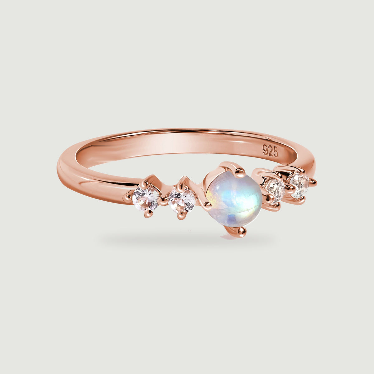 A rose gold ring with a round moonstone and four clear side stones rests on a light gray surface.