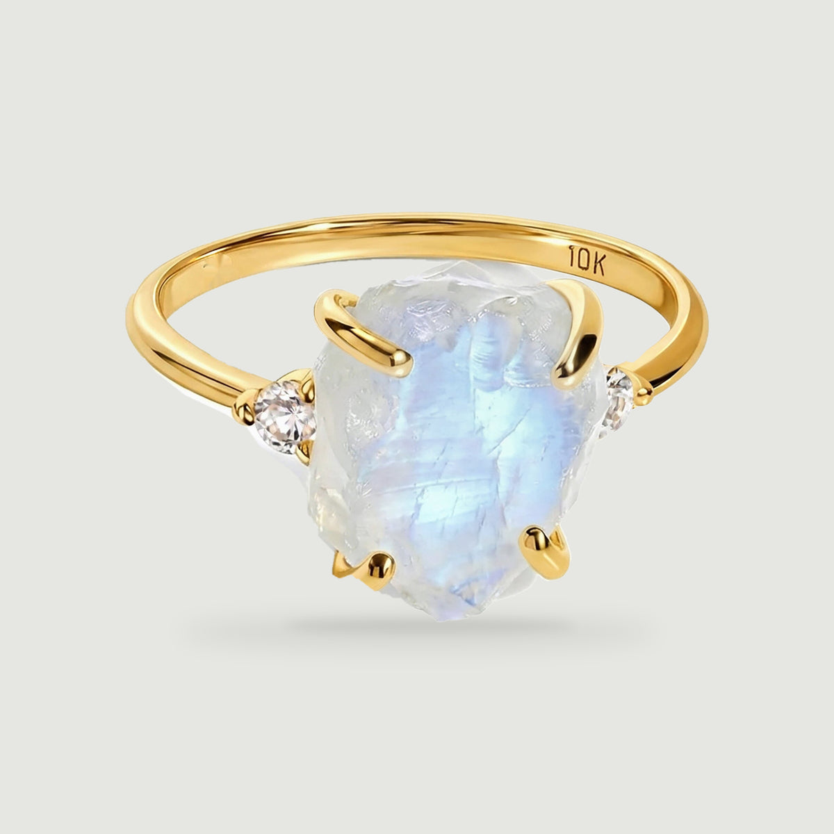 A yellow gold ring holds a rough pale moonstone between two round diamond accents on a light gray surface.