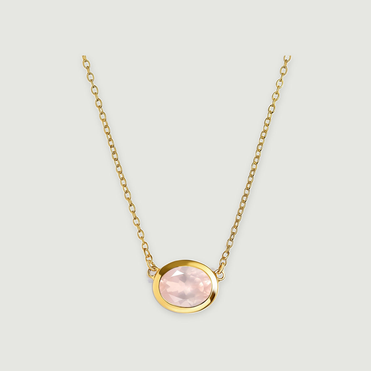 A yellow gold colored bezel surrounds a pale pink oval stone beneath two lengths of matching link chain on light gray.