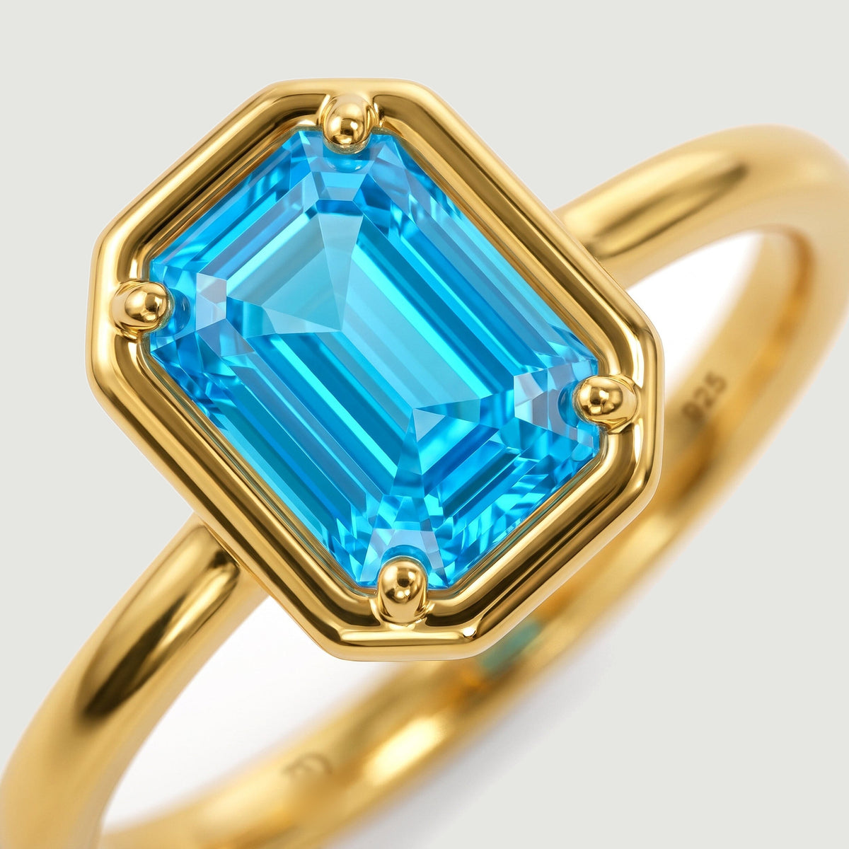 A blue rectangular gemstone fills a yellow gold setting with four rounded prongs.