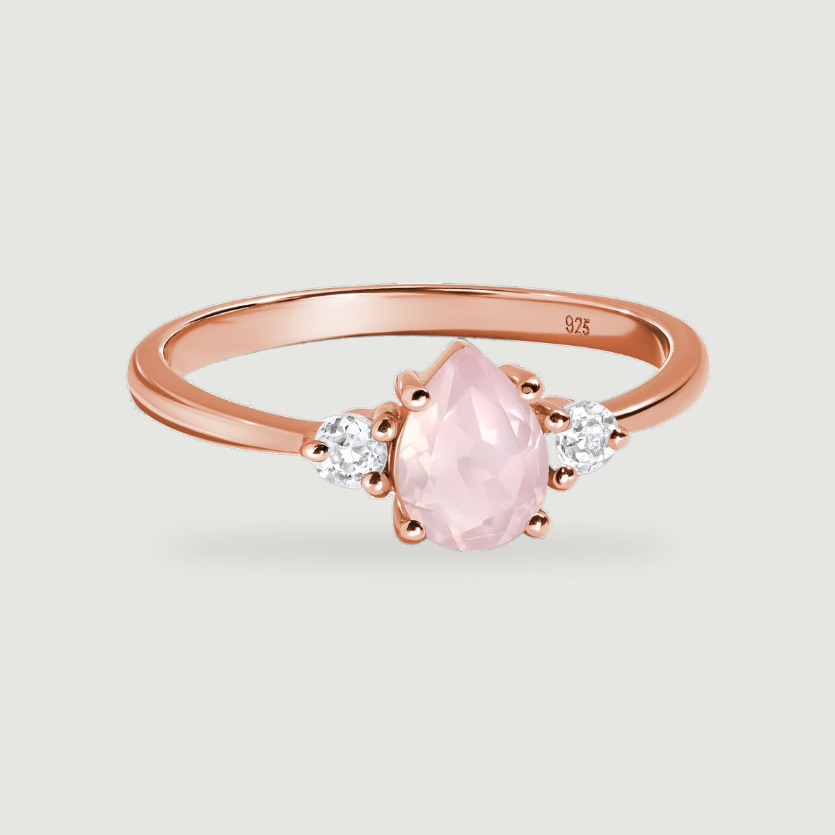 A rose gold ring with a pear-shaped pink gemstone and two round clear side stones rests on a light gray surface.