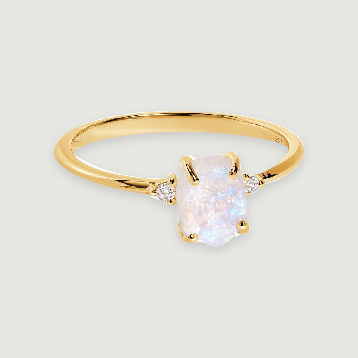 A yellow gold vermeil ring displays a rough moonstone between two small white zircon stones on a softly shadowed grey surface.