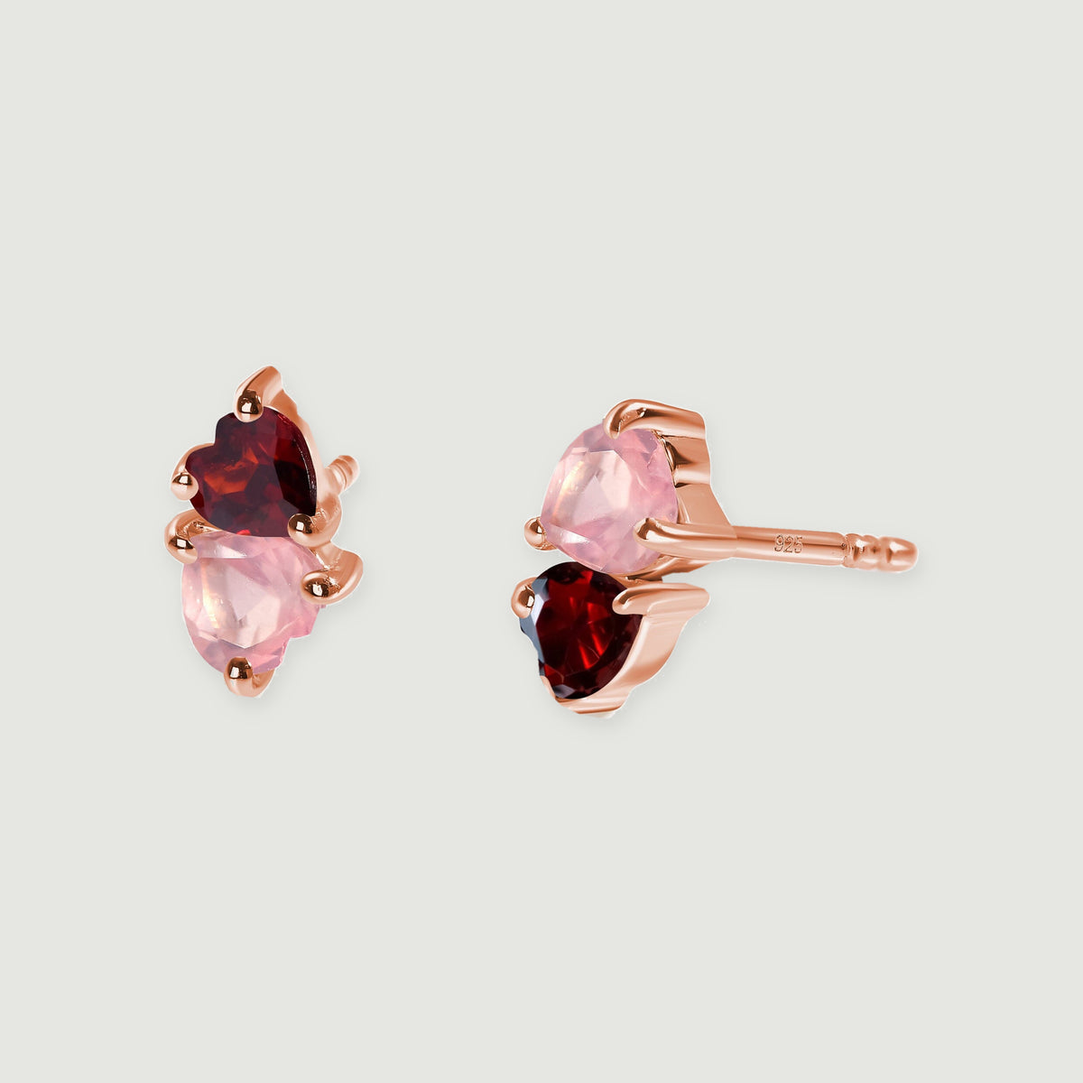 A pair of rose gold tone studs displays pale pink and deep red heart-shaped stones on a light gray surface.