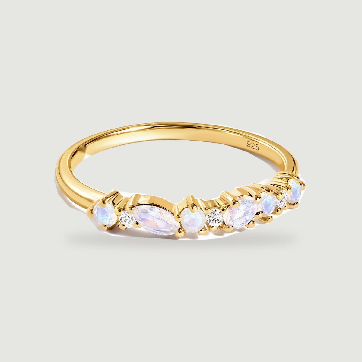 A yellow gold ring with mixed oval and round pale stones rests on a light gray surface.