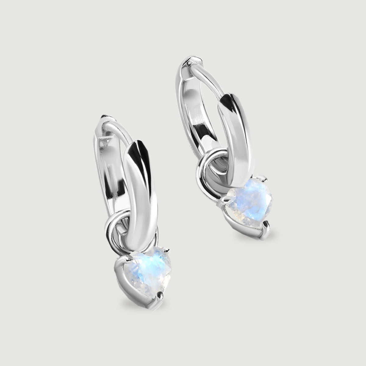 Two polished silver-tone hoop earrings hold heart-shaped moonstones on a light gray surface.