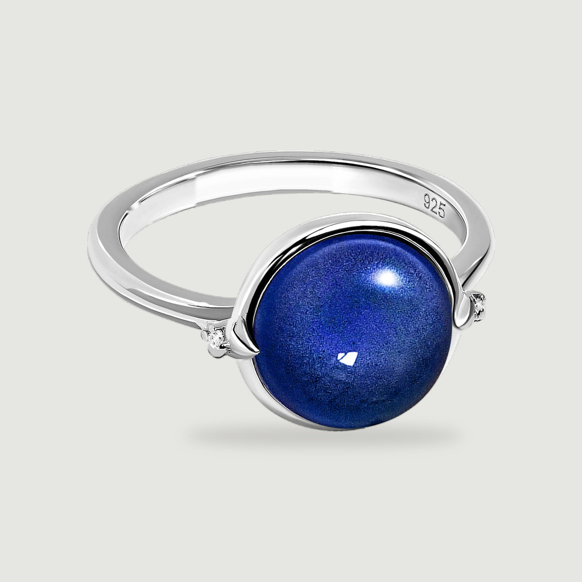 A polished silver ring holds a domed blue stone between two clear accents on a softly shaded grey background.