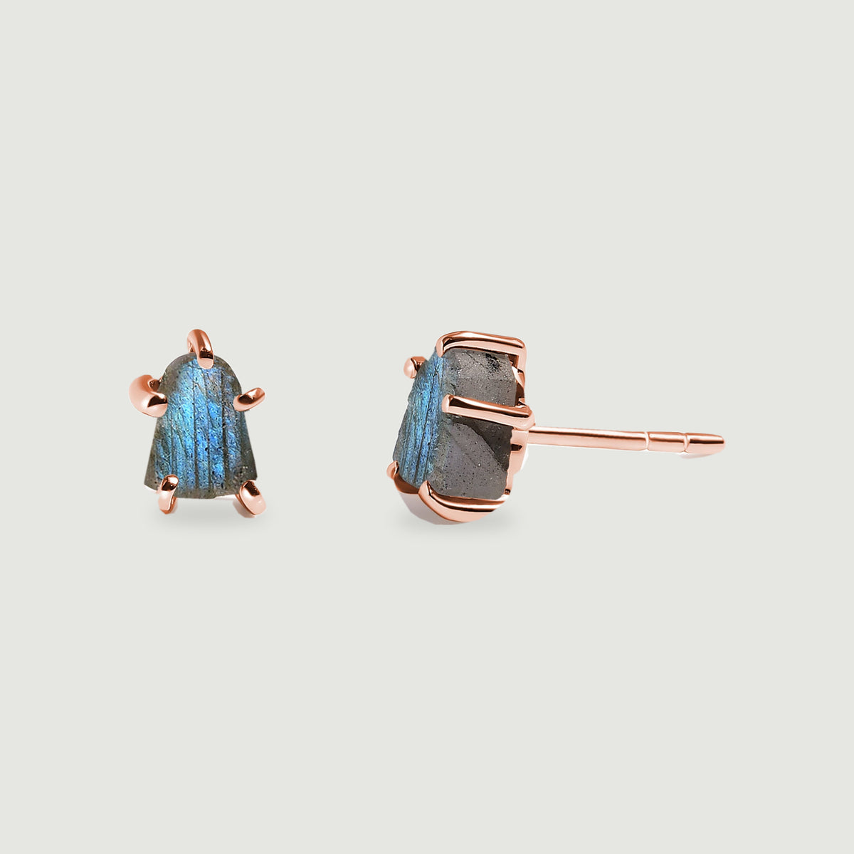 Rose gold-colored prongs hold two raw labradorite stones, with one stud facing forward and the other turned sideways on light gray.