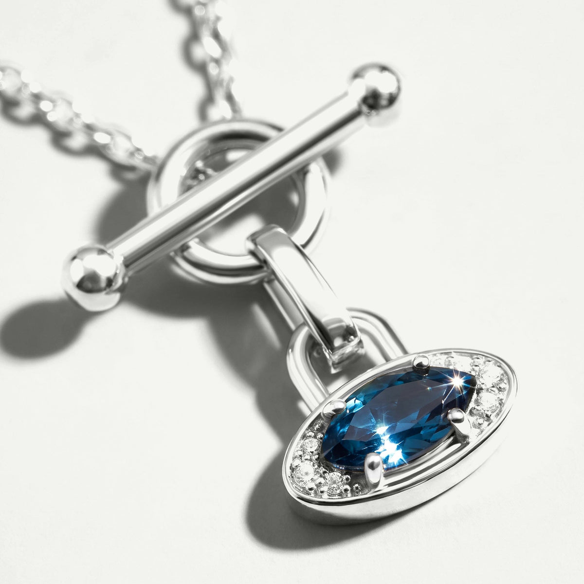 An angled close-up shows the silver pendant rim, deep blue gemstone and polished bar clasp on a light gray surface.