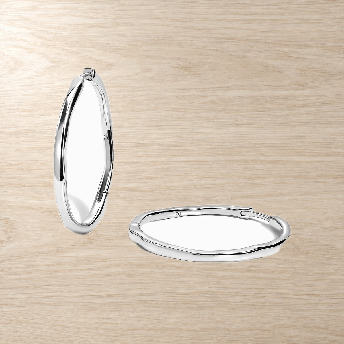 Pair of elongated wavy hoop earrings displayed on a light wood surface, with one hoop upright and one resting flat.