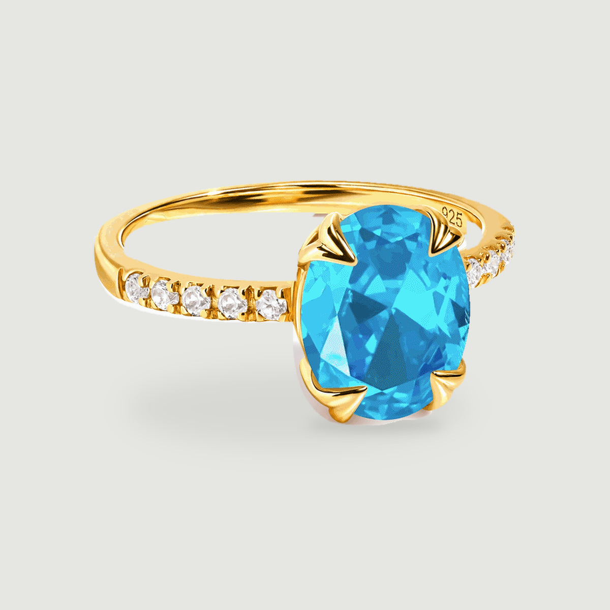 An oval blue gemstone sits in a yellow gold ring with four sculpted claws and a row of clear accent stones.