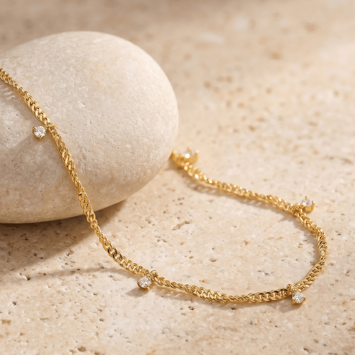 A yellow gold chain and small clear gemstone drops drape across a rounded stone and a cream tabletop.