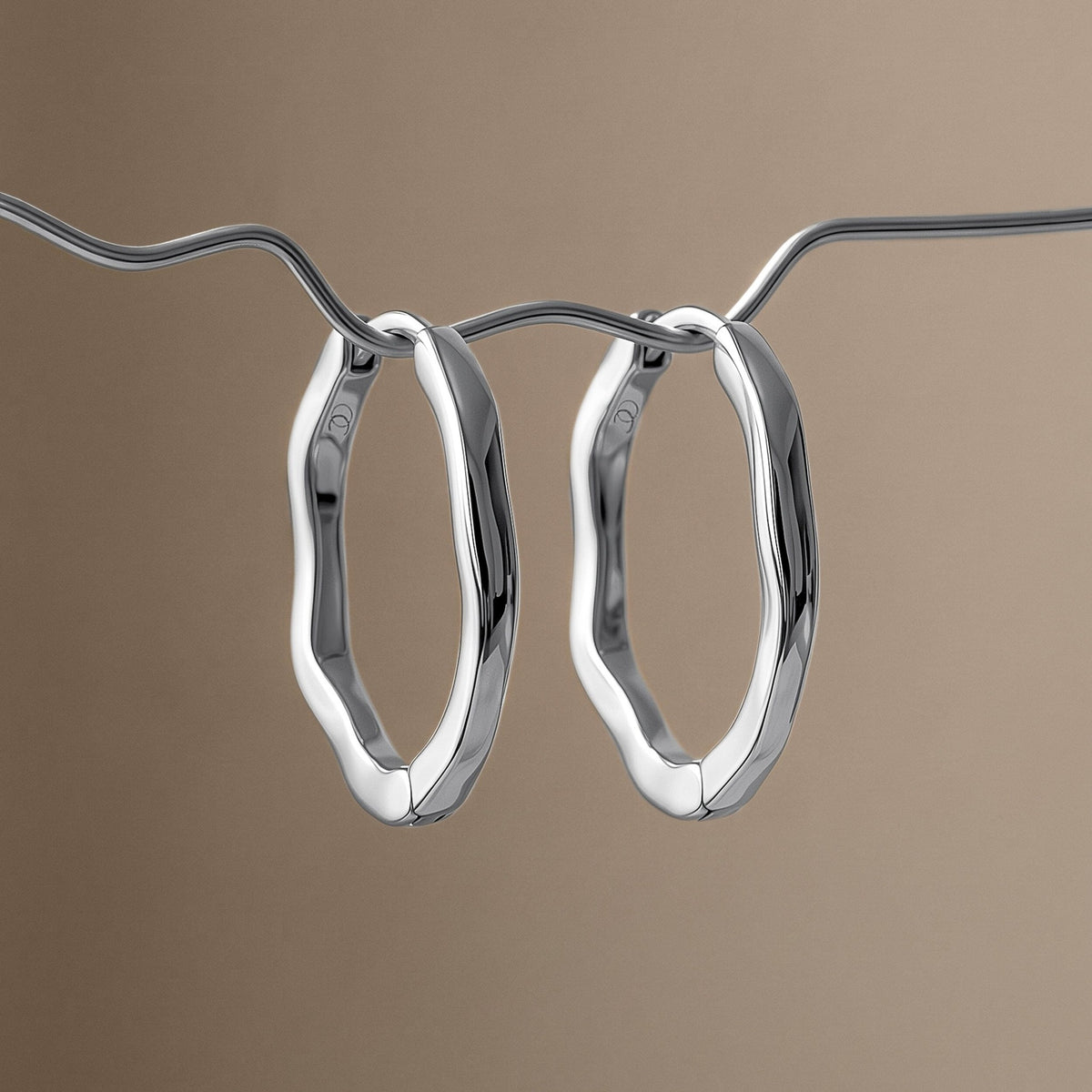 Pair of wavy oval hoops hanging from a curved chain against a warm taupe background.