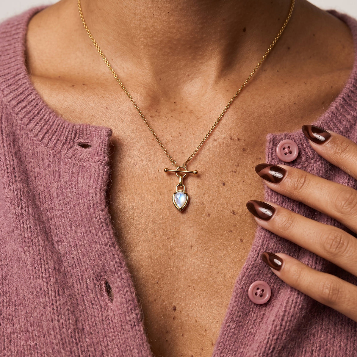 A hand with dark polished nails rests beside a pear-shaped moonstone toggle necklace on a mauve cardigan.