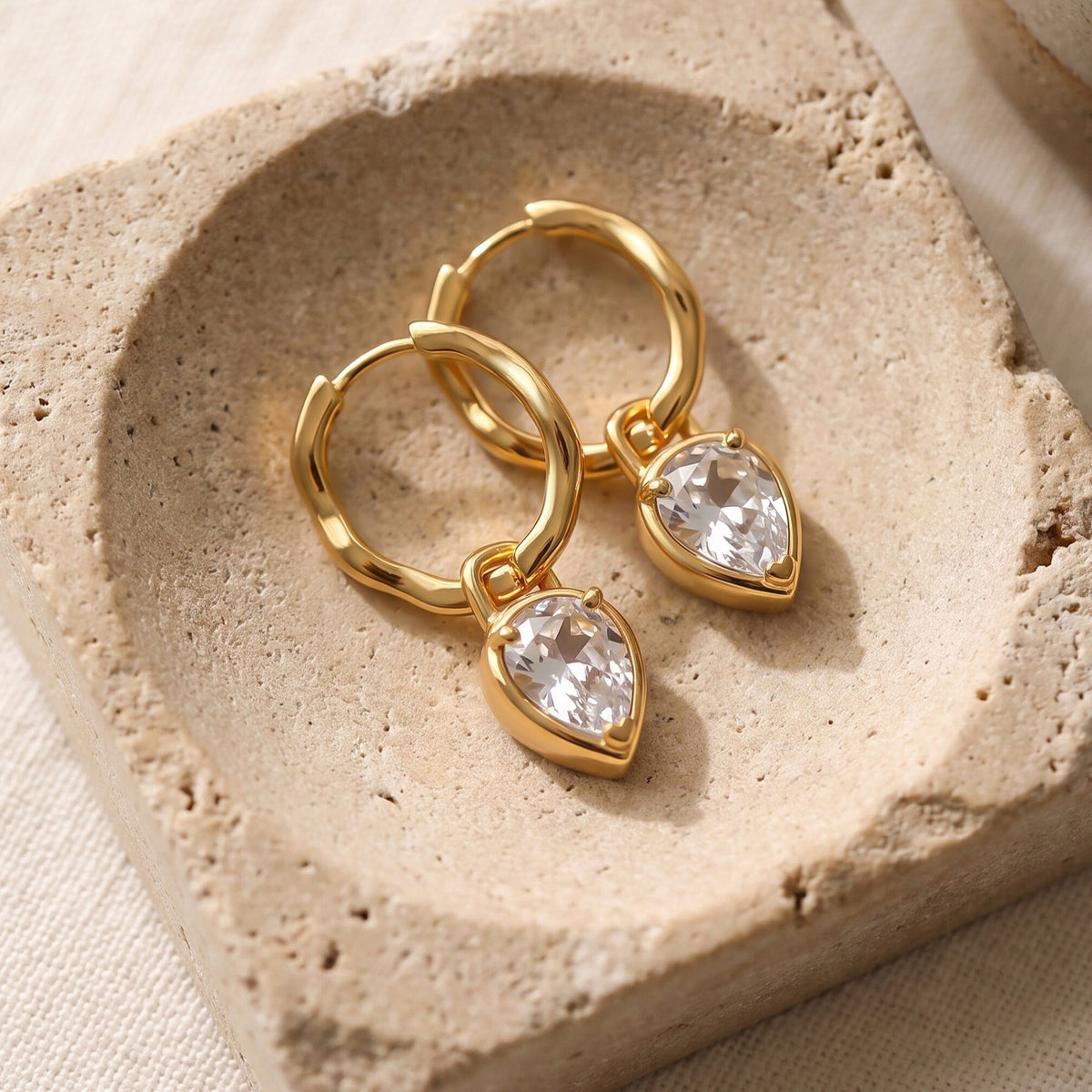 A pair of yellow-gold hoops with faceted clear drops lies on a beige stone dish.