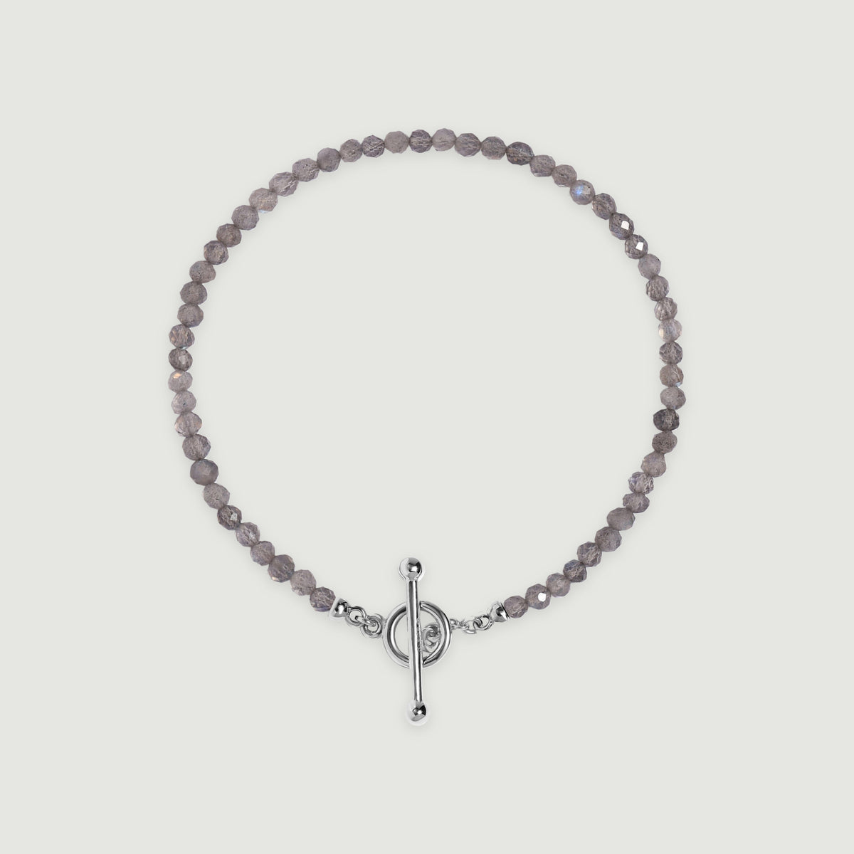 A complete grey labradorite bead bracelet with a polished silver ring and bar clasp lies centered on a light grey surface.