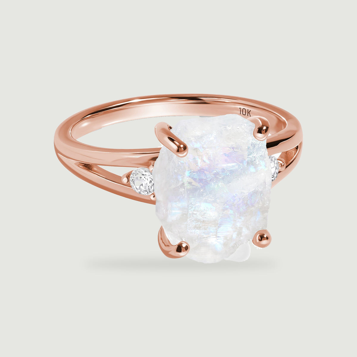 A rose gold ring with a rough white moonstone and two round clear side stones rests on a light gray background.