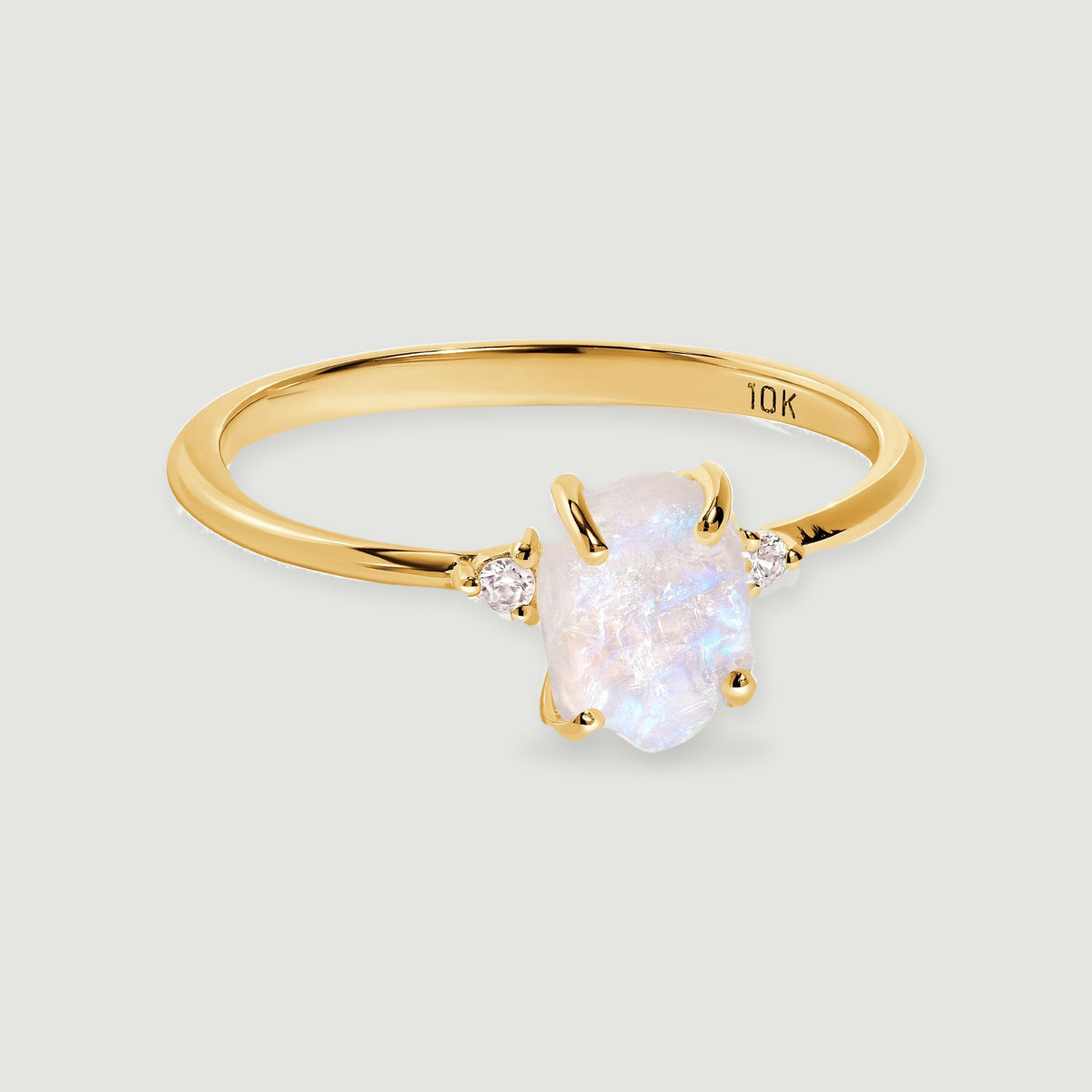 A yellow gold ring with a raw pale moonstone and two round diamond accents rests on a light gray surface.