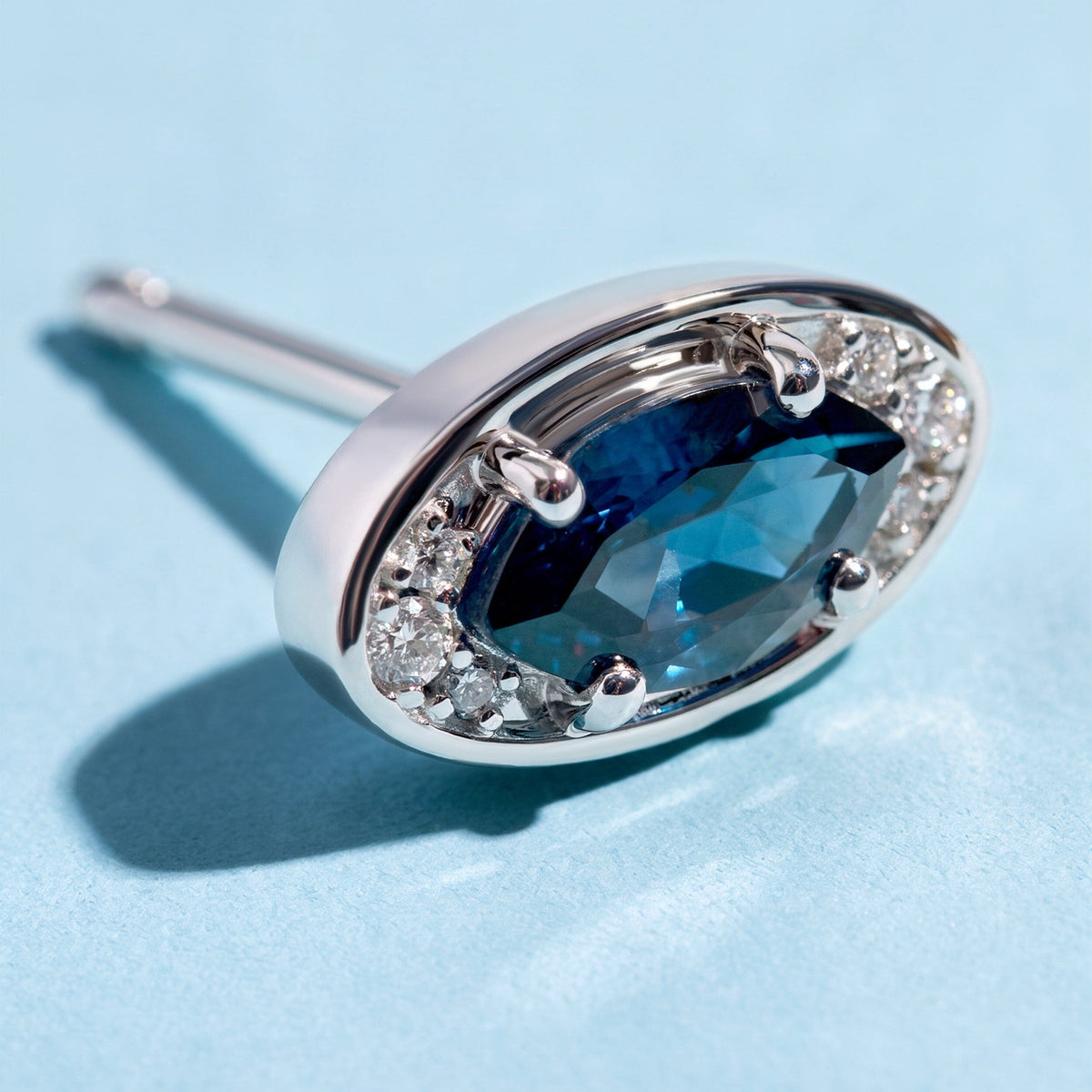 A close view of the silver-tone earring reveals four rounded prongs and clear stones beside a faceted blue center.