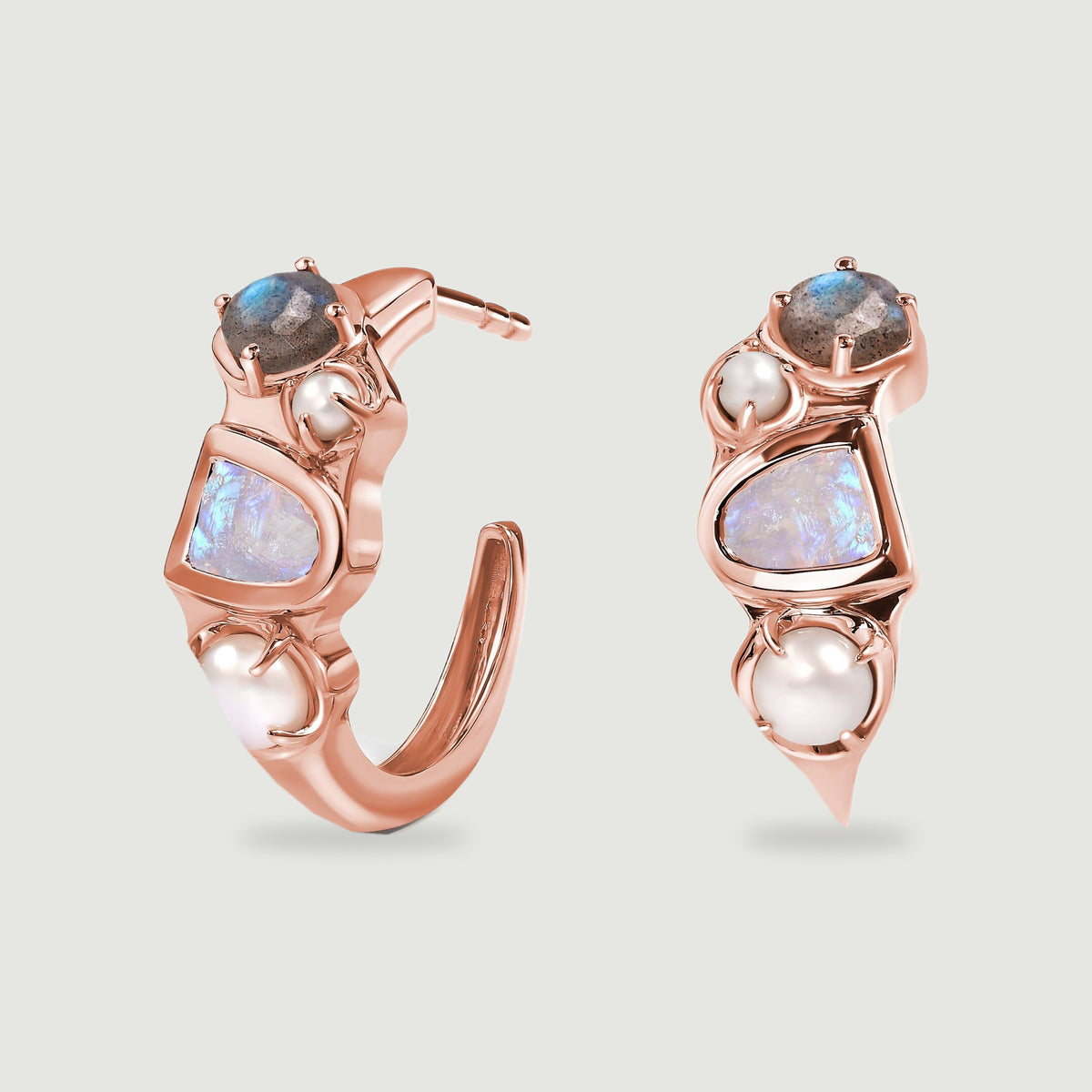 Two rose-gold-tone hoop earrings show polished curves, pearl accents and pale iridescent stones on a gray surface.