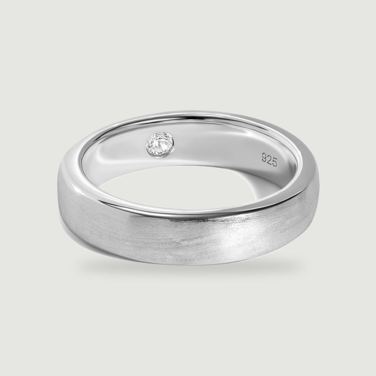 A brushed silver ring with a polished rim and a small clear stone inside rests on a light gray surface.