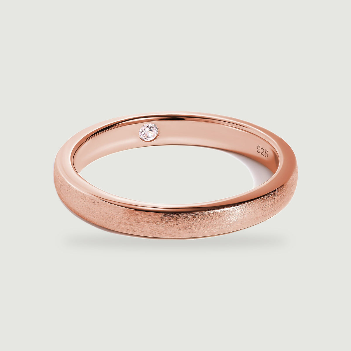A rose gold ring rests at an angle with a brushed band and a clear stone set into its inner surface.