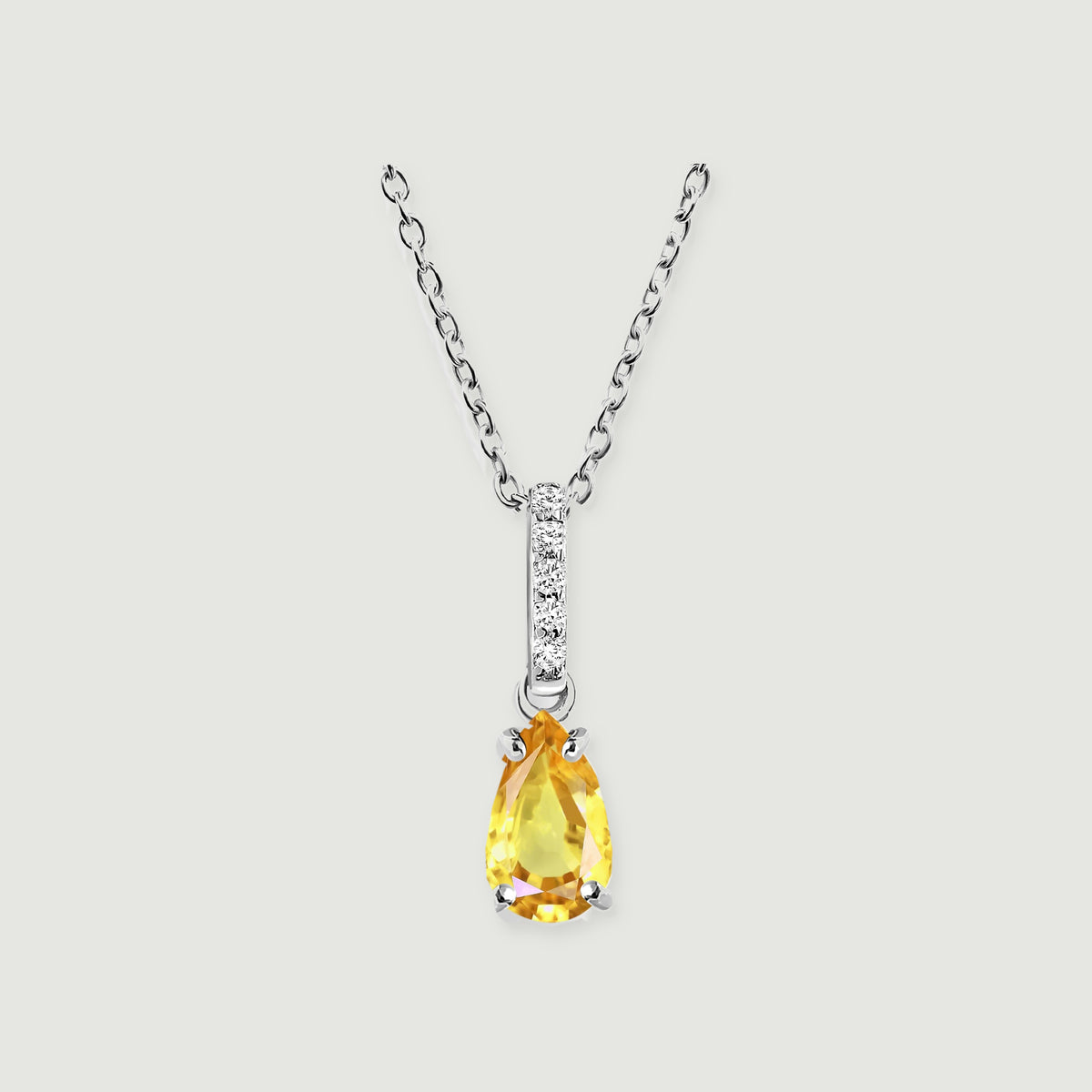 A silver necklace displays a yellow pear-shaped pendant beneath five round clear stones on a pale gray background.
