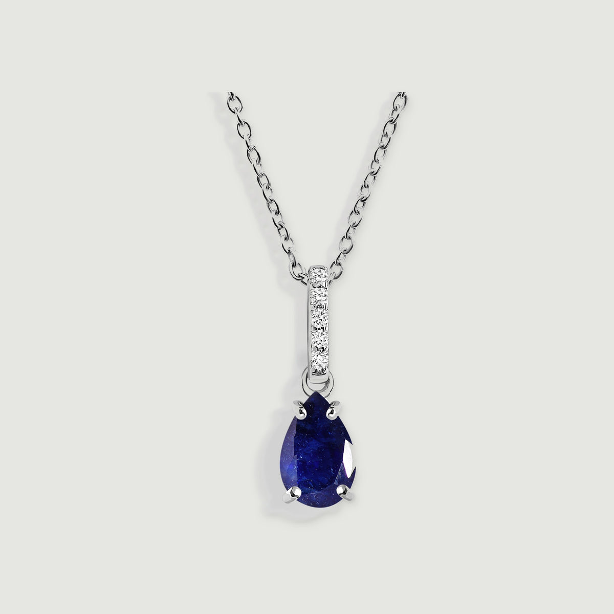 A silver link chain holds a blue pear-shaped pendant beneath a vertical row of five clear stones on a pale gray background.
