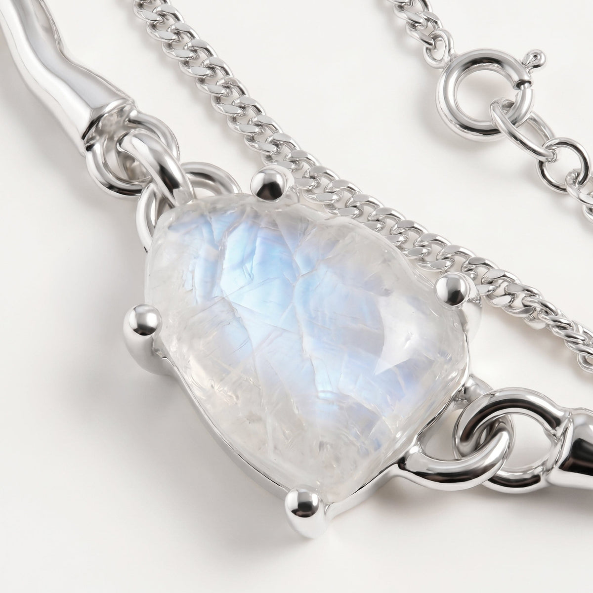 A close view shows blue reflections inside the pale moonstone beside silver prongs, a polished link and a fine chain on a clean tabletop.