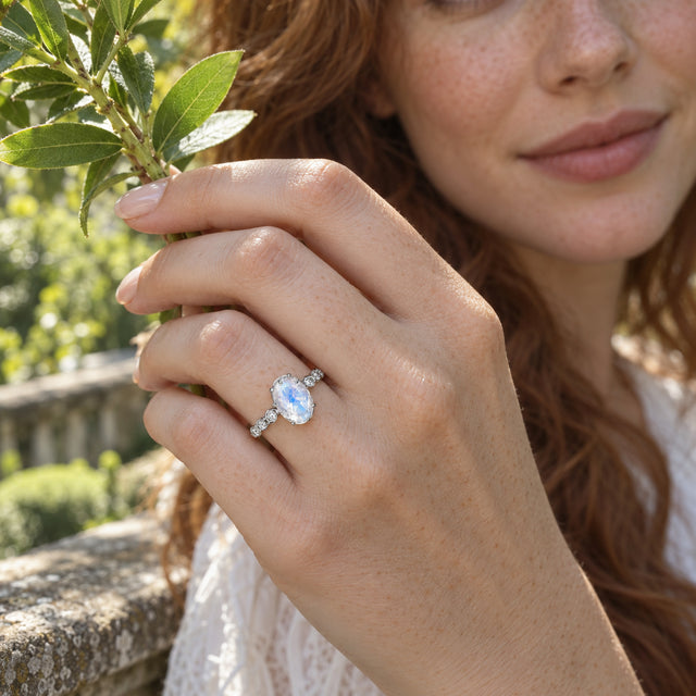 A white gold moonstone ring is worn on a freckled hand holding a leafy stem beside a garden terrace.