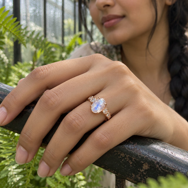 A rose gold ring with an oval moonstone and round diamond accents is worn on a hand resting on a greenhouse railing.