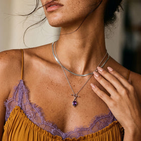 Neckline portrait shows the purple teardrop necklace layered with another chain and touched by a hand.