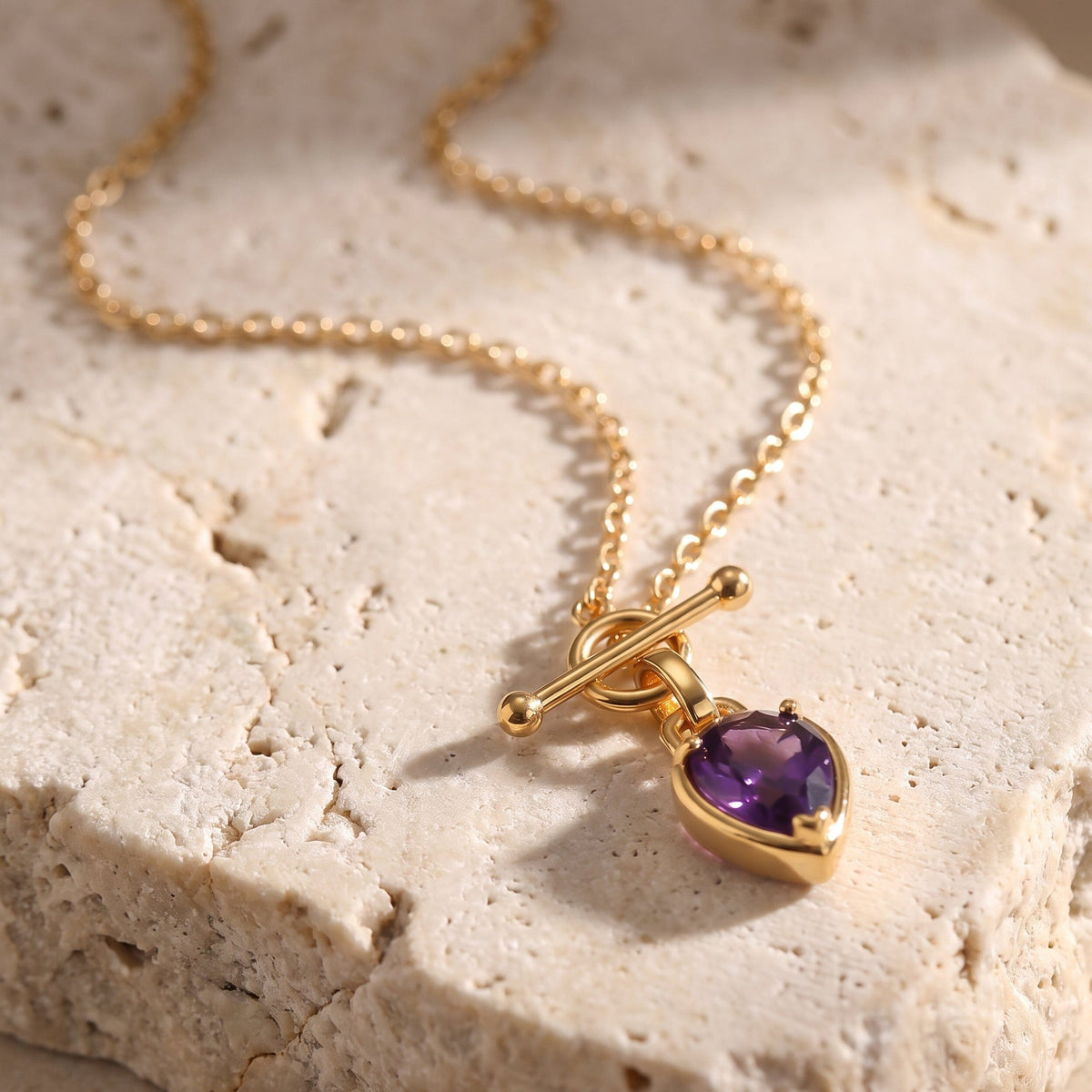 Pendant necklace rests on pale stone, showing a bar clasp and faceted purple rounded gemstone.