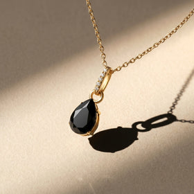 A yellow gold vermeil chain and black pear-shaped pendant appear above a warm stone tabletop in sunlight.