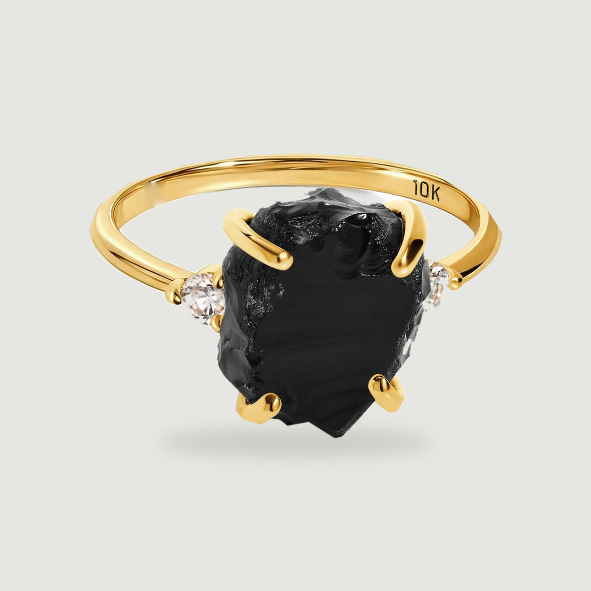 A yellow gold ring holds a rough black stone between two small round clear stones on a light gray background.