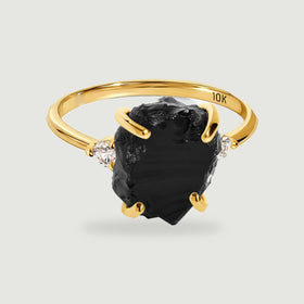 A yellow gold ring holds a rough black stone between two small round clear stones on a light gray background.