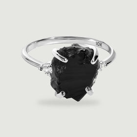 A white gold ring with an irregular black center stone and two clear side stones rests above a soft shadow.