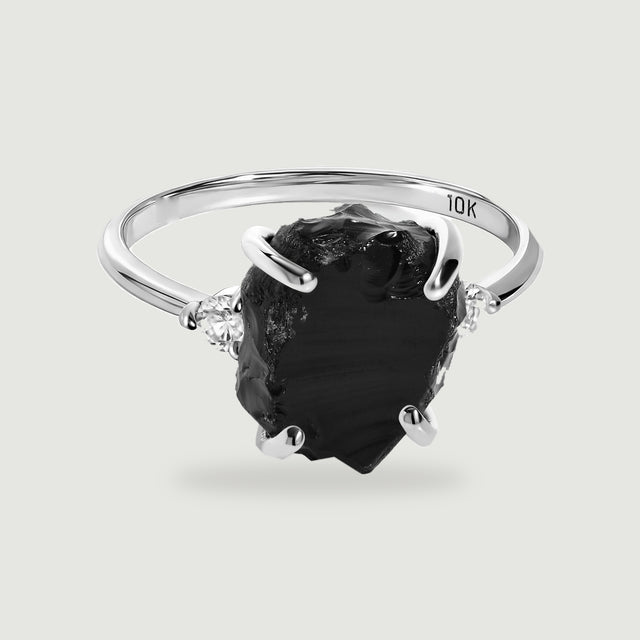A white gold ring with an irregular black center stone and two clear side stones rests above a soft shadow.