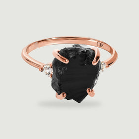 A rose gold ring shows a dark textured center stone, curved prongs, and two round clear accents.