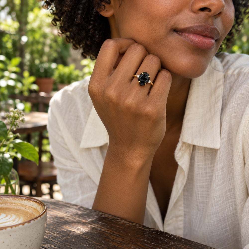 A rough black-stone ring with a yellow gold band is worn on a hand held beside the face at a leafy cafe.