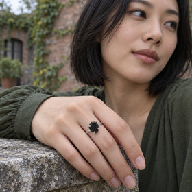 A white gold ring with a rough black center is worn on a hand resting on a weathered stone ledge.