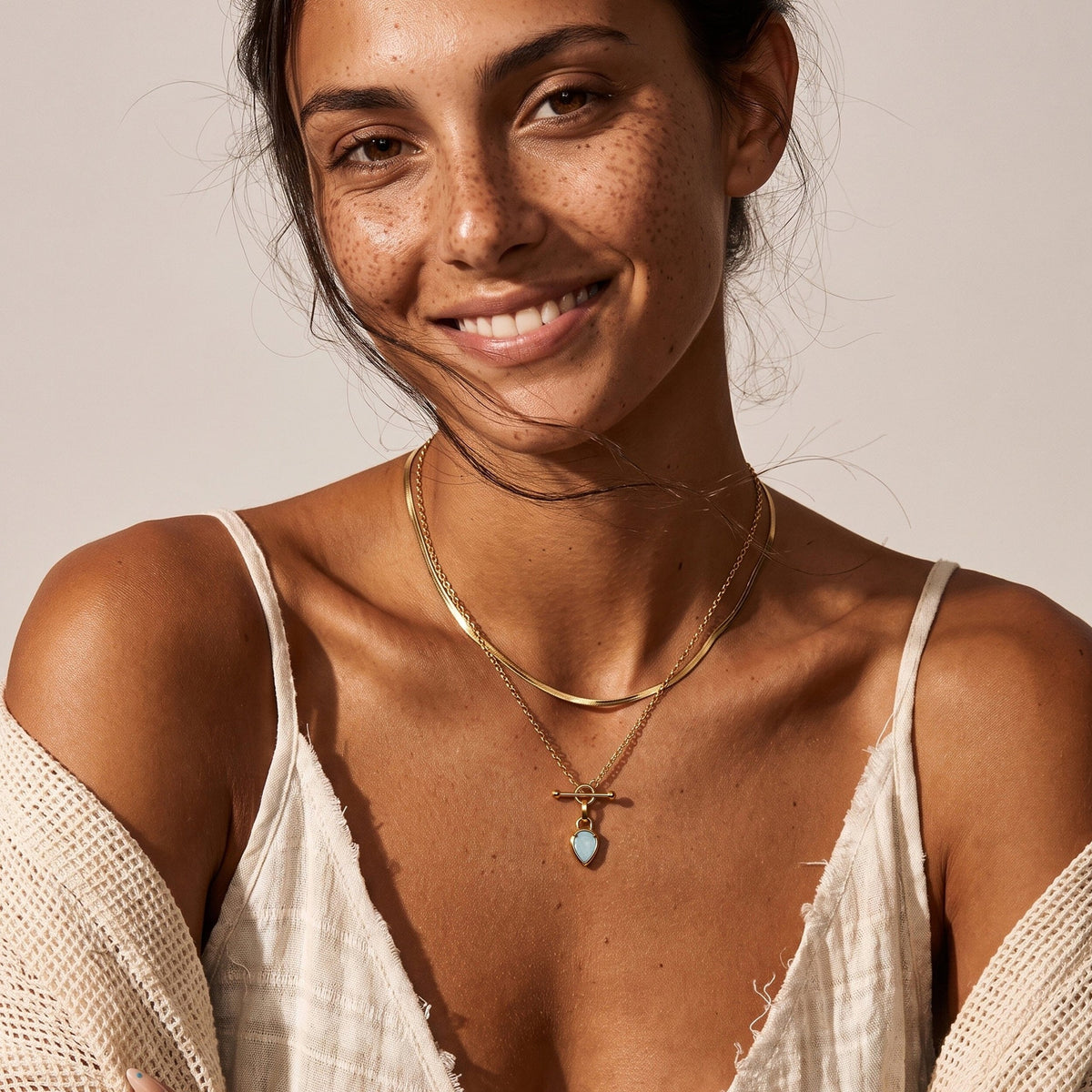 Smiling freckled woman wears the blue pear-shaped pendant necklace with layered chains and a white strappy top.