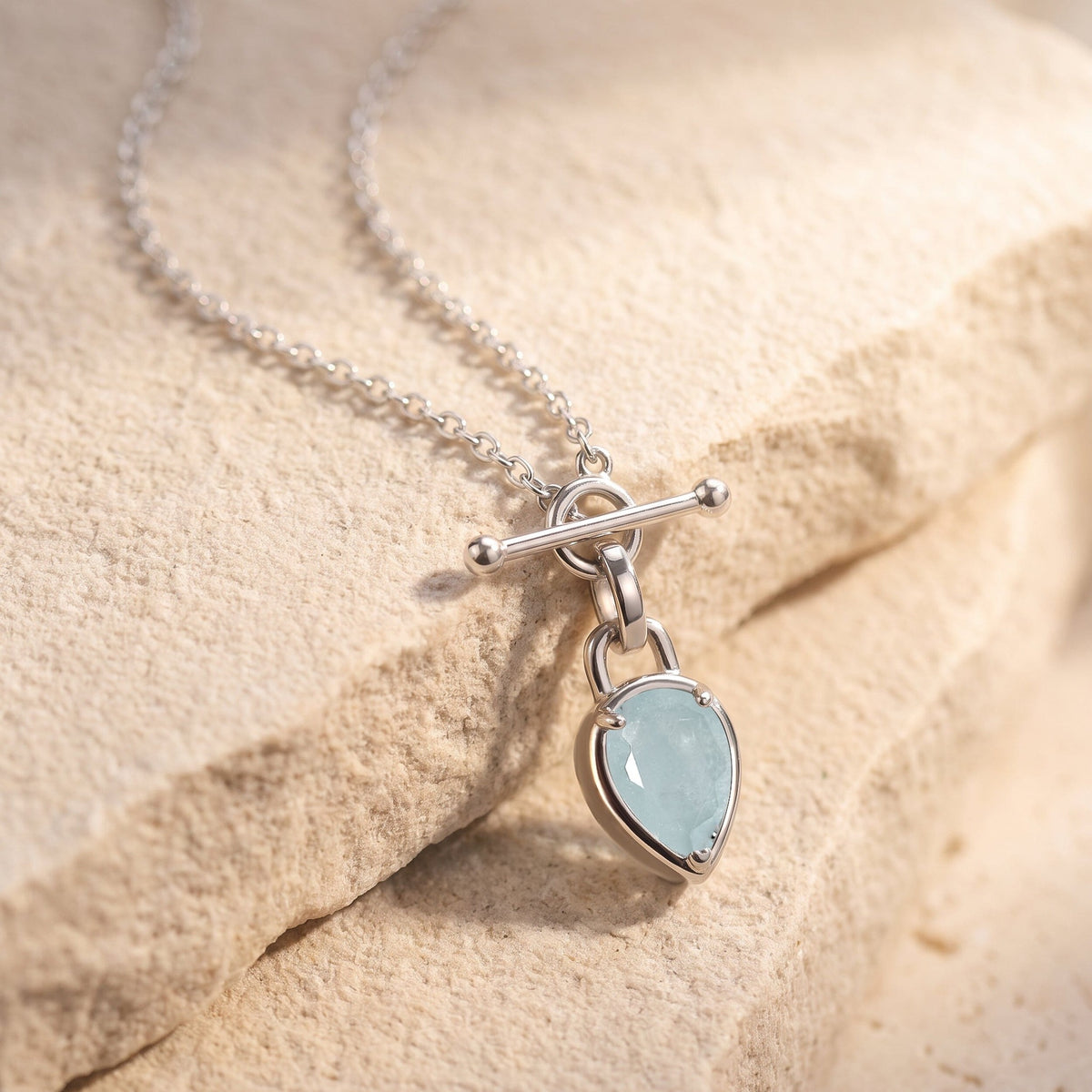 Pear-shaped pale blue pendant necklace draped across a textured beige stone surface in soft light.