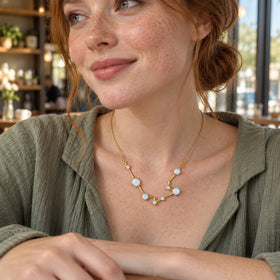 A freckled woman in a green blouse wears the gold constellation necklace beside a cafe window.