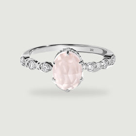 A white gold ring with an oval pale pink stone and three round diamonds on each shoulder rests on a light gray surface.