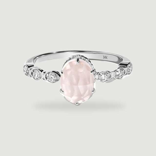 A white gold ring with an oval pale pink stone and three round diamonds on each shoulder rests on a light gray surface.