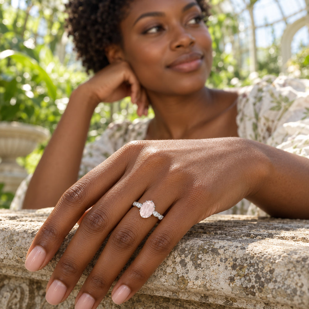 A white gold pink-stone ring is worn on a woman's hand resting on a stone bench with green foliage behind her.