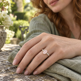 A yellow gold ring with an oval pink stone is shown on a freckled hand resting on sage linen at a garden table.