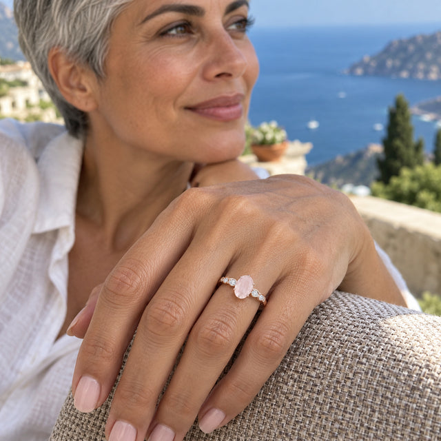A rose gold pink-stone ring with bright diamond accents sits on a woman's hand above a woven chair arm overlooking blue water.